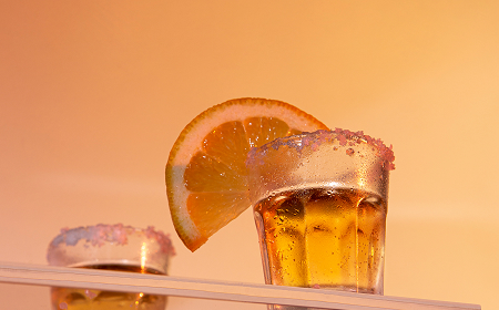 Стопка с янтарным напитком, украшенная долькой апельсина и сахарной каймой, на тёплом фоне